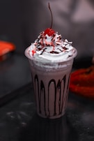 A creamy dessert served in a transparent cup, topped with whipped cream, a drizzle of chocolate syrup, and a cherry on top. The cup itself has vertical streaks of chocolate decorating its sides.