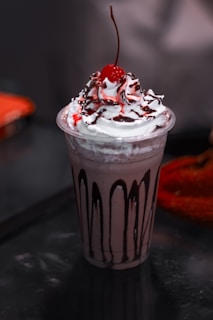 A creamy dessert served in a transparent cup, topped with whipped cream, a drizzle of chocolate syrup, and a cherry on top. The cup itself has vertical streaks of chocolate decorating its sides.