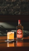 A bottle of Olmeca Reposado Tequila sits on a wooden table next to a cocktail garnished with citrus slices. The shadows and lighting suggest a warm, relaxed atmosphere.