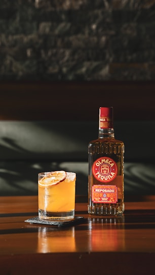 A bottle of Olmeca Reposado Tequila sits on a wooden table next to a cocktail garnished with citrus slices. The shadows and lighting suggest a warm, relaxed atmosphere.