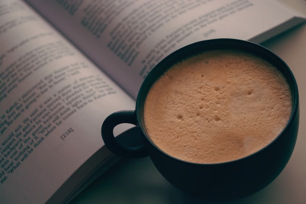 An open book with visible text lies on a flat surface next to a dark-colored mug filled with frothy coffee. The focus is on the mug and the page of the book, giving a cozy and inviting atmosphere.