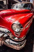 A close-up view of a vintage car with a bright red glossy finish featuring chrome detailing. The focus is on the front fender and headlight with reflections visible in the polished surfaces. The background includes an urban street setting with blurred buildings.