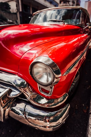 A close-up view of a vintage car with a bright red glossy finish featuring chrome detailing. The focus is on the front fender and headlight with reflections visible in the polished surfaces. The background includes an urban street setting with blurred buildings.