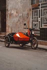 An orange motorcycle with a sidecar is parked on a street beside a weathered building wall. The wall features graffiti that reads 'LAURA TE AMO' with hearts around it. The building has a metal window grille and worn plaster.