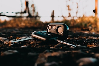 A set of car keys and a key fob are lying on a gravelly surface outdoors. The background is slightly blurred, highlighting the keys in the foreground. The lighting gives a warm, golden hue, suggesting a sunset or early morning setting.