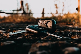 A set of car keys and a key fob are lying on a gravelly surface outdoors. The background is slightly blurred, highlighting the keys in the foreground. The lighting gives a warm, golden hue, suggesting a sunset or early morning setting.