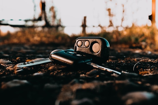 A set of car keys and a key fob are lying on a gravelly surface outdoors. The background is slightly blurred, highlighting the keys in the foreground. The lighting gives a warm, golden hue, suggesting a sunset or early morning setting.