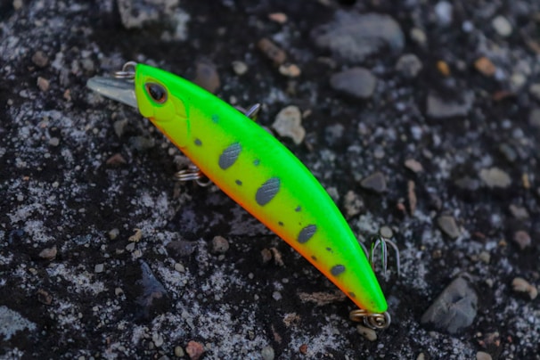 A bright and colorful fishing lure rests on a textured, rocky surface. The lure is predominantly neon green with orange highlights and black spots, equipped with metal hooks.