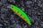 A bright and colorful fishing lure rests on a textured, rocky surface. The lure is predominantly neon green with orange highlights and black spots, equipped with metal hooks.