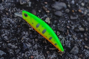 A bright and colorful fishing lure rests on a textured, rocky surface. The lure is predominantly neon green with orange highlights and black spots, equipped with metal hooks.
