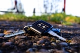 A close-up view of a set of car keys on a cobblestone surface with a rural, out-of-focus background featuring green grass and some blurred vertical structures.