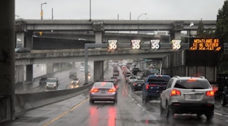A busy highway with heavy traffic during rainy weather. Overhead signs indicate a speed limit of 30, and there is an LED board displaying information about road closures. Multiple cars are moving slowly, and the road surface is wet and reflective.