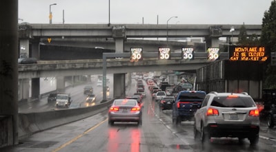 A busy highway with heavy traffic during rainy weather. Overhead signs indicate a speed limit of 30, and there is an LED board displaying information about road closures. Multiple cars are moving slowly, and the road surface is wet and reflective.