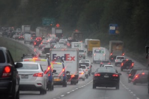 Heavy traffic on a multi-lane highway under overcast skies. Several cars and delivery trucks, including FedEx vehicles, are visible. Brake lights are on, indicating slow movement. Trees line the sides of the highway, and road signs are in the background.
