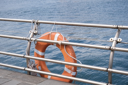 An orange lifebuoy is attached to a metal railing using ropes. The background consists of a body of calm blue water, suggesting a waterfront or seaside location. The scene conveys a sense of safety and preparedness.