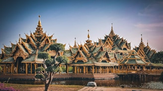 An intricately designed structure with ornate, pointed roofs and elaborate details stands beside a reflective body of water. The architecture features an array of peaks and tiers, typical of traditional Southeast Asian styles. Surrounding the structure are well-maintained trees and lush greenery.