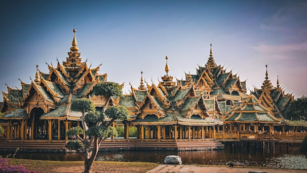 An intricately designed structure with ornate, pointed roofs and elaborate details stands beside a reflective body of water. The architecture features an array of peaks and tiers, typical of traditional Southeast Asian styles. Surrounding the structure are well-maintained trees and lush greenery.