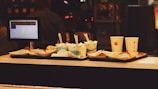 A fast food counter with trays holding several paper cups, some with straws, and small food items wrapped in paper. A screen displaying a menu is visible on the left side, suggesting a self-service or order kiosk. The background is dimly lit, giving it a cozy ambiance.