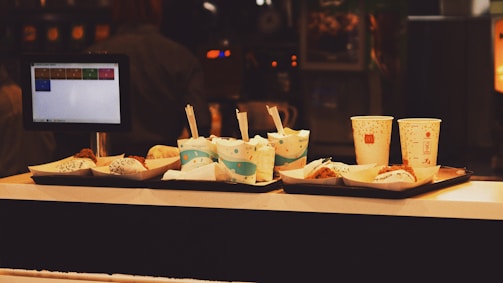 A fast food counter with trays holding several paper cups, some with straws, and small food items wrapped in paper. A screen displaying a menu is visible on the left side, suggesting a self-service or order kiosk. The background is dimly lit, giving it a cozy ambiance.
