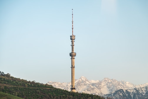 A tall communication tower stands prominently against a backdrop of snow-capped mountains. The landscape includes a green, tree-covered slope in the foreground, with a clear blue sky above.