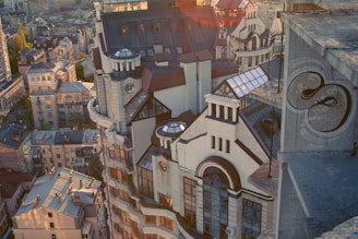 An aerial view of an urban area featuring a prominent, ornate building with a mix of modern and classical architectural elements, including large windows, geometric shapes, and decorative rooftops. The cityscape extends into the background with numerous residential buildings. Sunlight casts warm highlights and soft shadows across the scene.