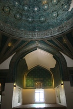 A richly decorated interior features an intricate geometric pattern design on the ceiling, with a dominant teal color and white starbursts. The walls are adorned with similar detailed patterns and a pointed arch frames a window allowing soft light to enter the room.