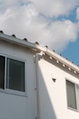 A section of a building’s exterior wall is shown, featuring a smooth white finish with two rectangular windows. The building has a light gray gutter running vertically. Above, the roof has a simple design with small triangular sections. The sky is mostly clear, with soft, fluffy white clouds scattered across a light blue background.
