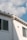 A section of a building’s exterior wall is shown, featuring a smooth white finish with two rectangular windows. The building has a light gray gutter running vertically. Above, the roof has a simple design with small triangular sections. The sky is mostly clear, with soft, fluffy white clouds scattered across a light blue background.