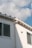 A section of a building’s exterior wall is shown, featuring a smooth white finish with two rectangular windows. The building has a light gray gutter running vertically. Above, the roof has a simple design with small triangular sections. The sky is mostly clear, with soft, fluffy white clouds scattered across a light blue background.