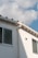 A section of a building’s exterior wall is shown, featuring a smooth white finish with two rectangular windows. The building has a light gray gutter running vertically. Above, the roof has a simple design with small triangular sections. The sky is mostly clear, with soft, fluffy white clouds scattered across a light blue background.