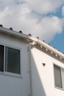 A section of a building&rsquo;s exterior wall is shown, featuring a smooth white finish with two rectangular windows. The building has a light gray gutter running vertically. Above, the roof has a simple design with small triangular sections. The sky is mostly clear, with soft, fluffy white clouds scattered across a light blue background.