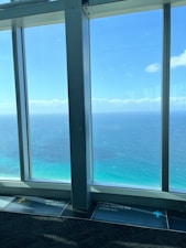 Large windows overlook a vast expanse of ocean and sky, with different shades of blue blending into the horizon. The ocean has a gradient from turquoise near the shore to a deeper blue further out. There are markings on the floor indicating distances to various cities such as New York and Los Angeles.