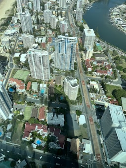 An aerial view of an urban area with numerous high-rise buildings, streets, and a river running alongside. The scene includes residential and commercial spaces, with some buildings surrounded by greenery and swimming pools. The layout of roads and intersections is clearly visible.