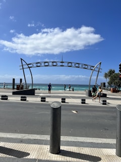 A coastal scene featuring a prominent archway labeled 'Surfers Paradise' with people walking along the sidewalk and beachgoers visible in the distance. The sky is bright and clear with a few clouds, and the ocean is a vivid blue.