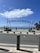 A coastal scene featuring a prominent archway labeled 'Surfers Paradise' with people walking along the sidewalk and beachgoers visible in the distance. The sky is bright and clear with a few clouds, and the ocean is a vivid blue.
