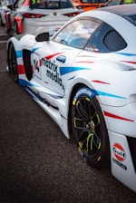 A white sports car with colorful blue and red streaks is parked on a tarmac. The car has various sponsor logos, including Michelin and Gulf, and the number 14 is prominently displayed along with the text 'matrix media'. Other race cars are visible in the background.