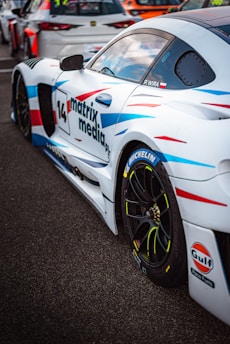 A white sports car with colorful blue and red streaks is parked on a tarmac. The car has various sponsor logos, including Michelin and Gulf, and the number 14 is prominently displayed along with the text 'matrix media'. Other race cars are visible in the background.