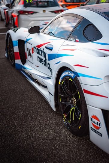A white sports car with colorful blue and red streaks is parked on a tarmac. The car has various sponsor logos, including Michelin and Gulf, and the number 14 is prominently displayed along with the text 'matrix media'. Other race cars are visible in the background.