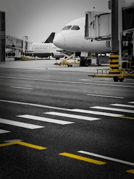 A commercial airplane is parked at an airport gate with jet bridges attached. The foreground shows a tarmac with white and yellow road markings. Another airplane can be seen in the background, and various airport equipment is present.