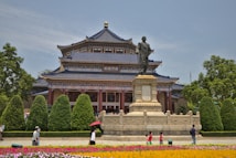 A traditional Chinese building with intricate architecture and a dark blue tiled roof stands prominently in a garden. In front of the structure, there is a large statue on a stone pedestal. The area is surrounded by neatly trimmed bushes and vibrant flowers. People are walking casually, some with umbrellas, adding to the lively atmosphere.