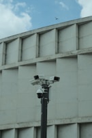 A group of security cameras mounted on a black pole stands in front of a geometric concrete building. The cameras are pointed in different directions, and a pigeon flies in the background against a partly cloudy sky.