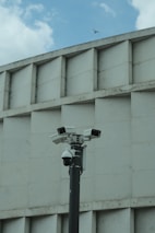 A group of security cameras mounted on a black pole stands in front of a geometric concrete building. The cameras are pointed in different directions, and a pigeon flies in the background against a partly cloudy sky.