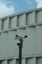 A group of security cameras mounted on a black pole stands in front of a geometric concrete building. The cameras are pointed in different directions, and a pigeon flies in the background against a partly cloudy sky.