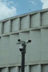 A group of security cameras mounted on a black pole stands in front of a geometric concrete building. The cameras are pointed in different directions, and a pigeon flies in the background against a partly cloudy sky.