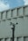A group of security cameras mounted on a black pole stands in front of a geometric concrete building. The cameras are pointed in different directions, and a pigeon flies in the background against a partly cloudy sky.