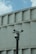 A group of security cameras mounted on a black pole stands in front of a geometric concrete building. The cameras are pointed in different directions, and a pigeon flies in the background against a partly cloudy sky.