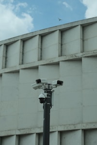 A group of security cameras mounted on a black pole stands in front of a geometric concrete building. The cameras are pointed in different directions, and a pigeon flies in the background against a partly cloudy sky.