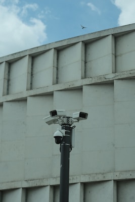 A group of security cameras mounted on a black pole stands in front of a geometric concrete building. The cameras are pointed in different directions, and a pigeon flies in the background against a partly cloudy sky.