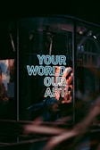 A storefront window displays a neon sign with the words 'Your World Our Art' in glowing white-blue light. The reflection of the street and surrounding buildings are visible in the glass, along with a faint outline of displayed items or artwork inside.