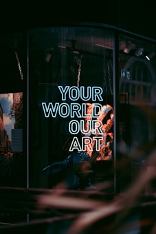 A storefront window displays a neon sign with the words 'Your World Our Art' in glowing white-blue light. The reflection of the street and surrounding buildings are visible in the glass, along with a faint outline of displayed items or artwork inside.